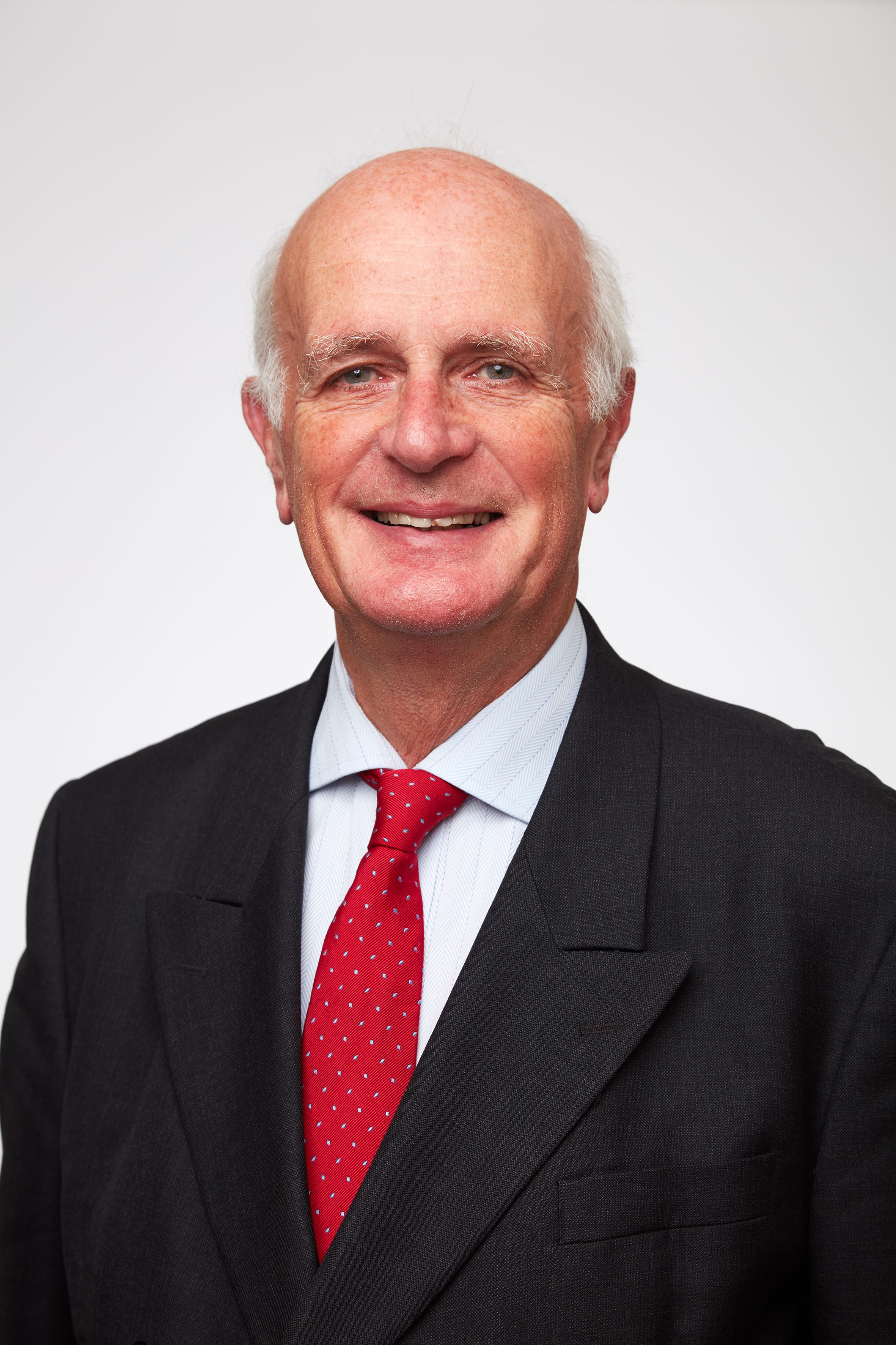 Mature man with white hair, wearing a dark suit and red patterned tie, smiling.